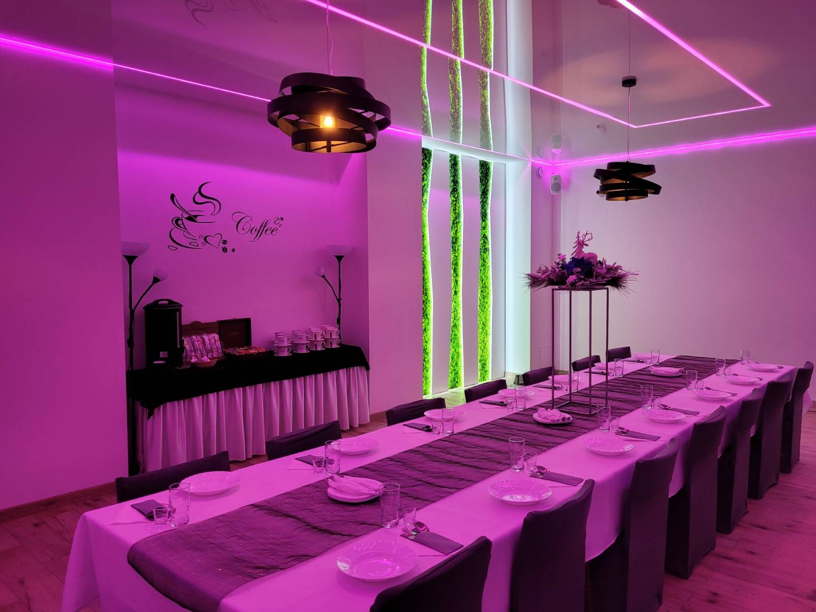 Elegancka sala bankietowa z długim stołem przygotowanym do przyjęcia, oświetlona neonowym, fioletowym światłem, z dekoracjami z mchu na ścianie i nowoczesnymi lampami.