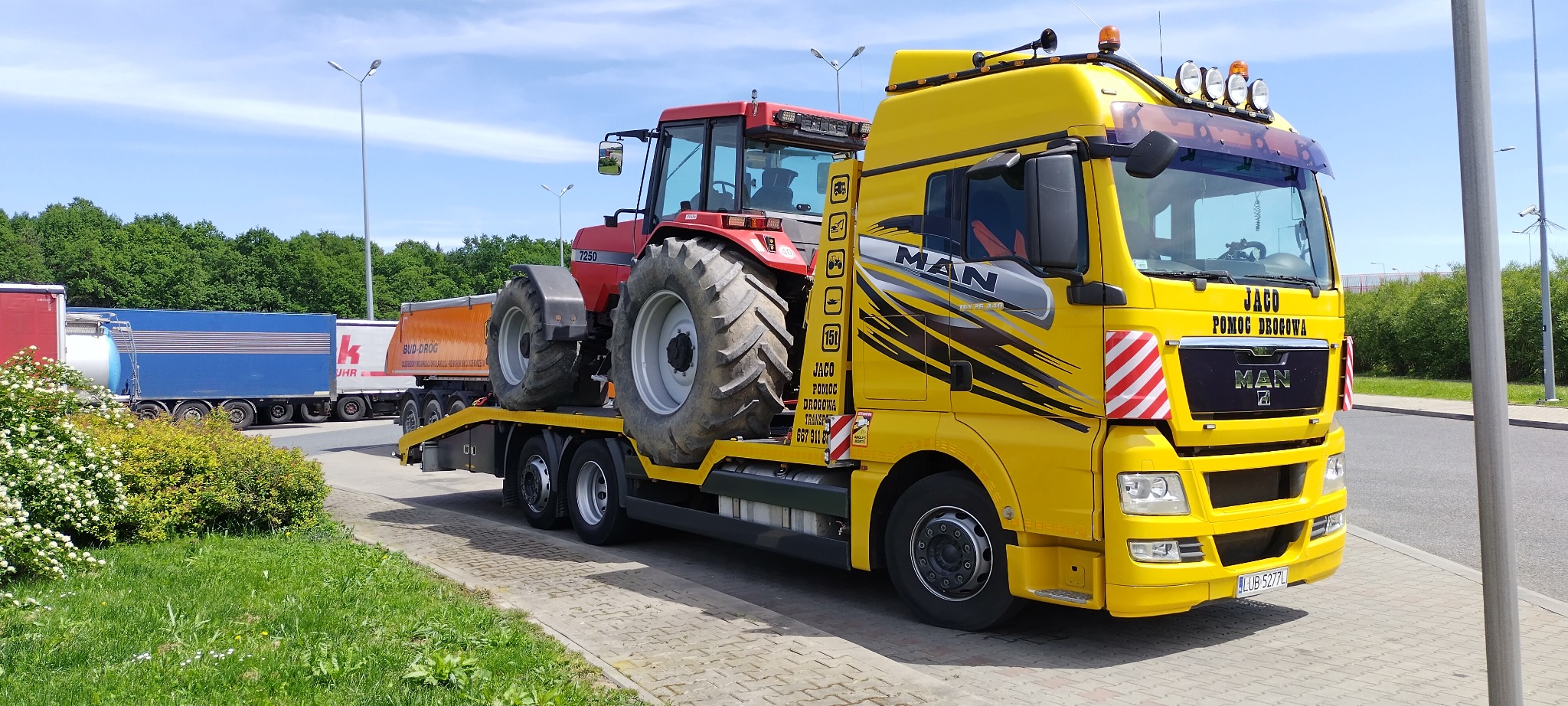 Żółta laweta MAN z czerwonym traktorem Case 7250 na platformie transportowej, widoczne logo firmy 'JACO Pomoc Drogowa Transport', w tle inne ciężarówki i zieleń.