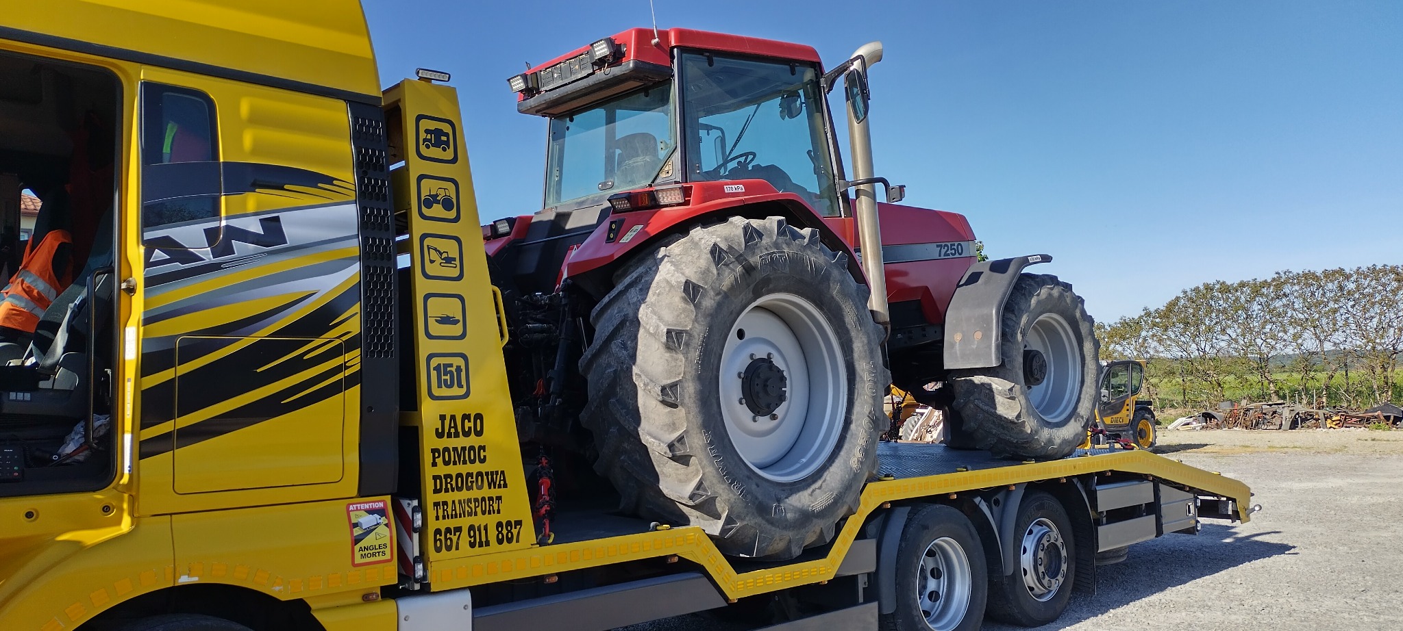 Czerwony traktor Case 7250 przewożony na żółtej lawecie pomocy drogowej MAN, widoczne oznaczenia firmy transportowej i numer telefonu, w tle krajobraz wiejski i inny sprzęt rolniczy.