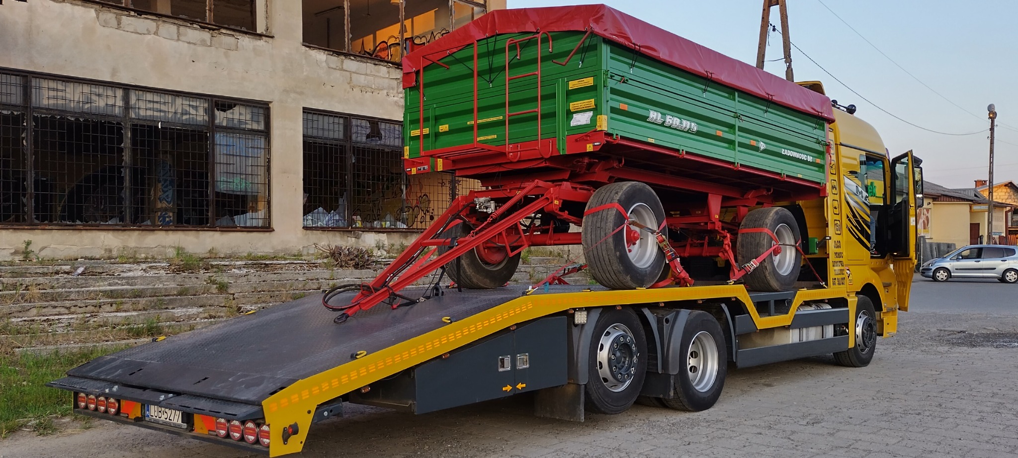 Żółta laweta przewozi zielono-czerwoną przyczepę rolniczą zabezpieczoną czerwonymi pasami transportowymi; w tle zrujnowany budynek.