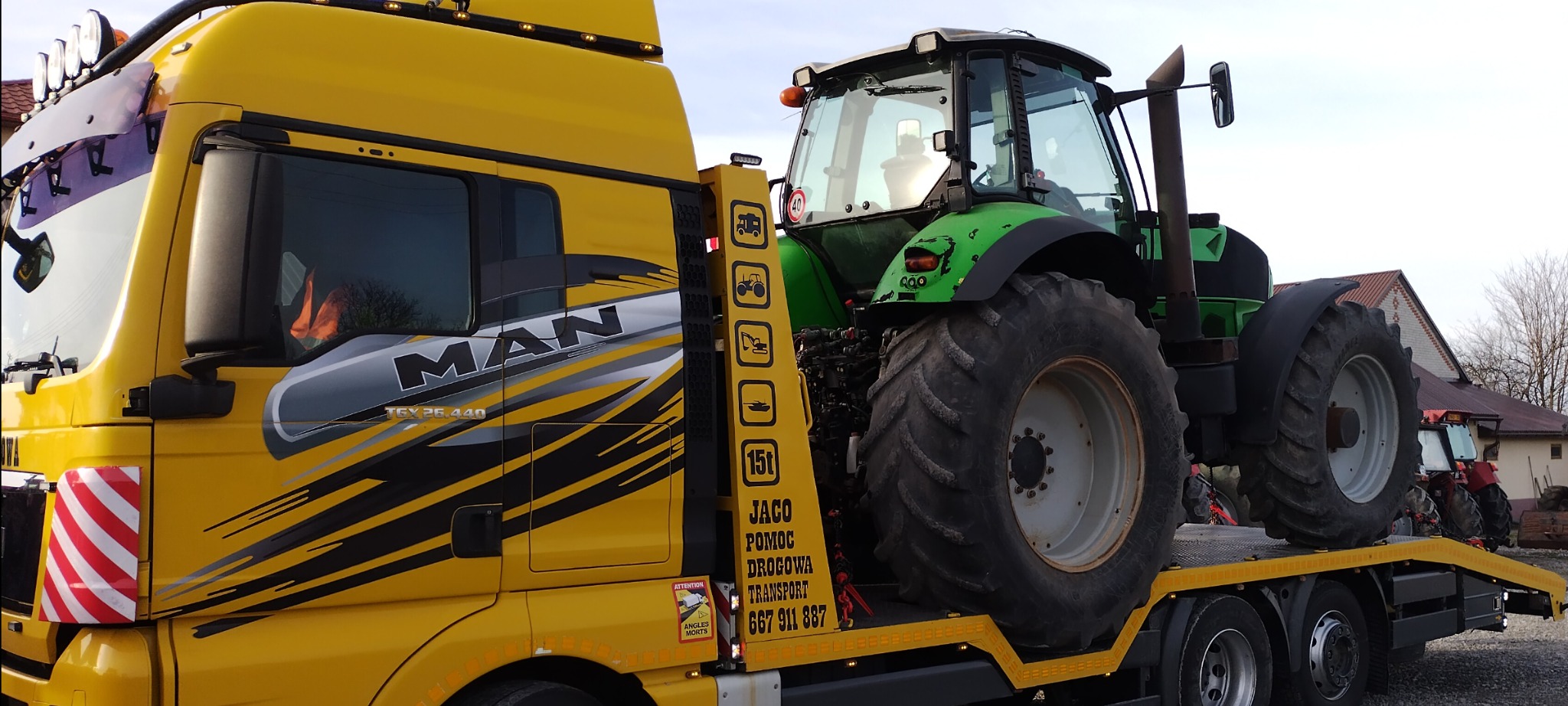 Żółta laweta MAN z zielonym traktorem na platformie, widoczne numery kontaktowe firmy transportowej i grafiki symbolizujące holowane pojazdy.