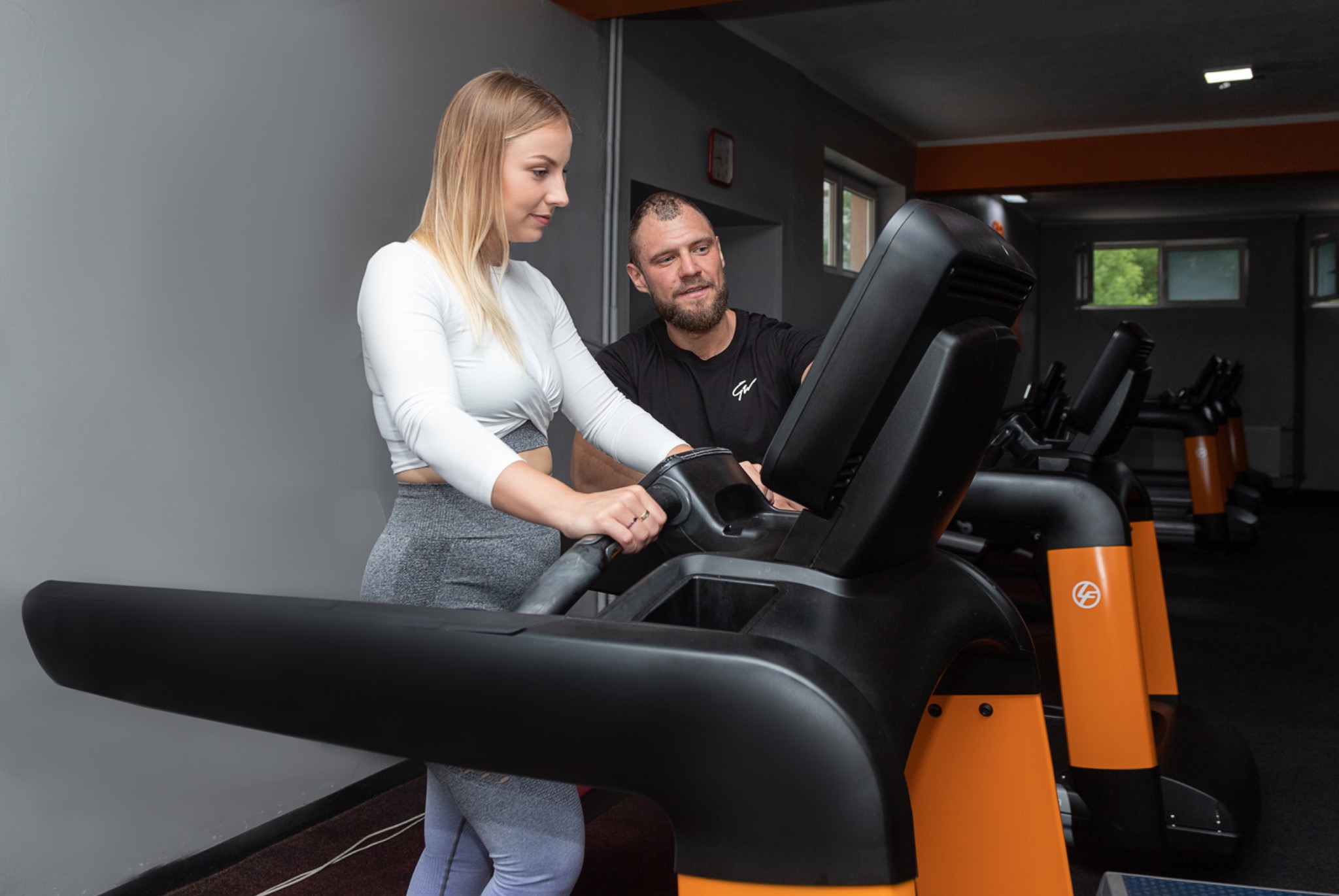 Kobieta na bieżni z instruktorem w tle, ćwicząca w jasnoszarym stroju sportowym z długim rękawem, skupiona na treningu, z widocznym pierścionkiem na palcu.