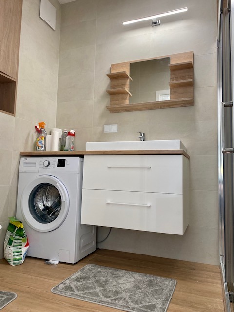 Nowoczesna łazienka z pralką obok białej szafki umywalkowej z drewnianym blatem, lustro z półkami w odcieniach drewna, beżowe płytki na ścianach i podłodze, dywanik w geometryczny wzór.