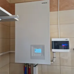 Biała pompa ciepła Hitachi zamontowana na ścianie z beżowych płytek, z widocznym panelem sterowania wskazującym godzinę 13:34 i datę 4 kwietnia 2023, oraz skrzynką elektryczną Noark obok.