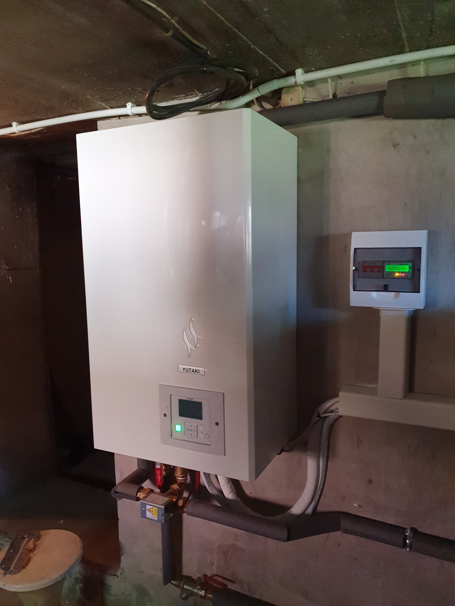 Biała pompa ciepła Yutaki zamontowana w pomieszczeniu technicznym, widoczne rury i skrzynka elektryczna z wyświetlaczem.