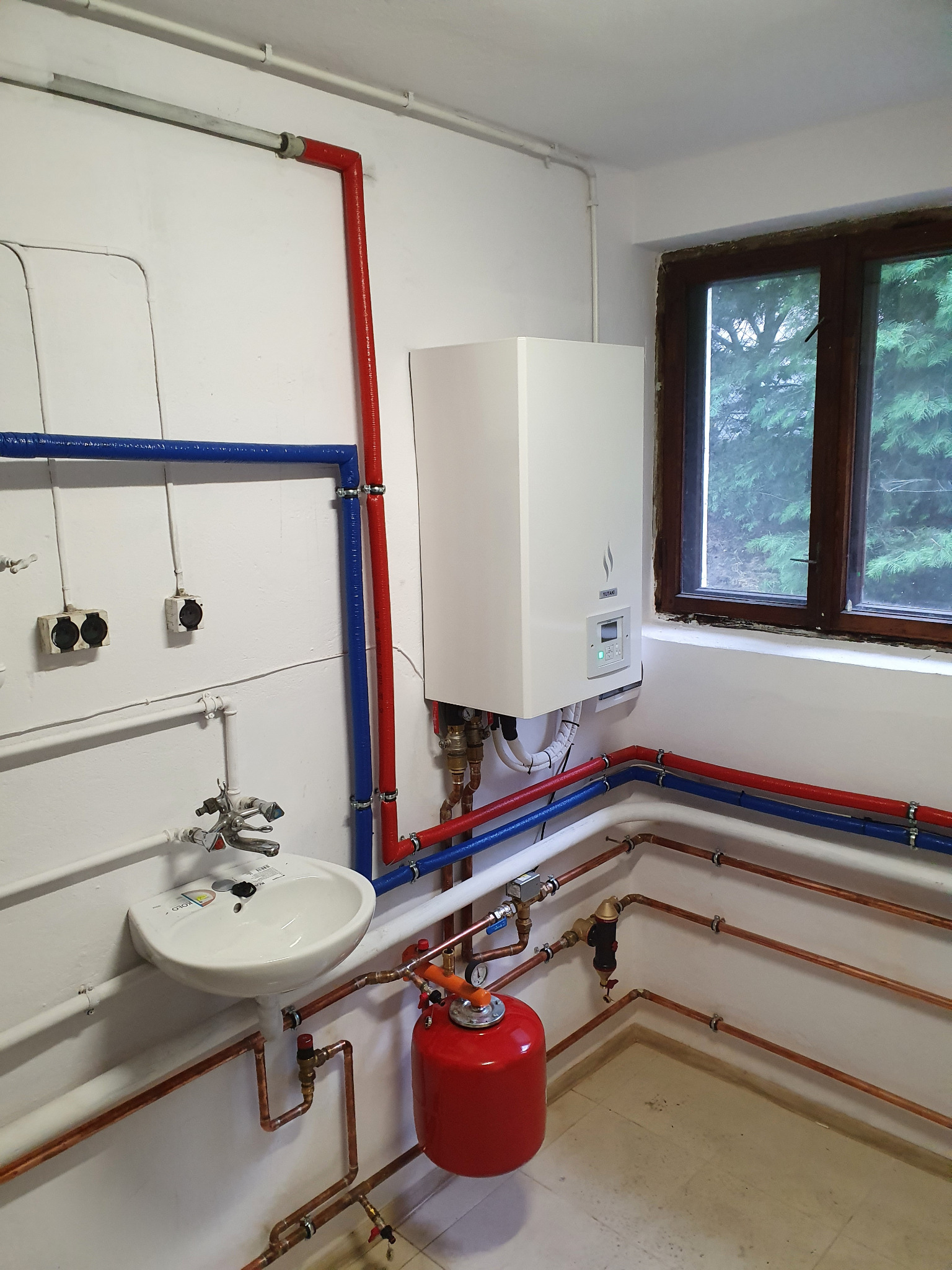 Wnętrze kotłowni z zamontowanym białym kotłem grzewczym Yutaki, widoczne rury miedziane, czerwone i niebieskie rury izolowane, biała umywalka oraz czerwony zbiornik wyrównawczy ciśnienia.