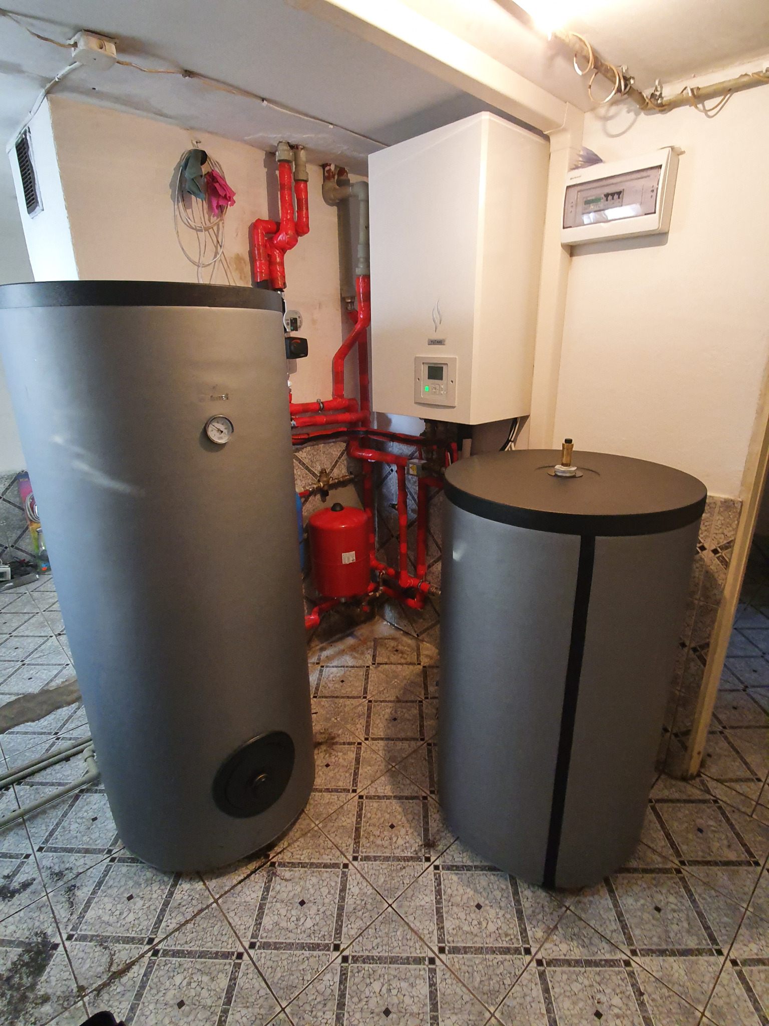 Dwa szare zasobniki ciepłej wody użytkowej w kotłowni, z widocznymi czerwonymi rurami instalacji grzewczej i białym piecem grzewczym na ścianie, na tle wzorzystej podłogi z płytek.