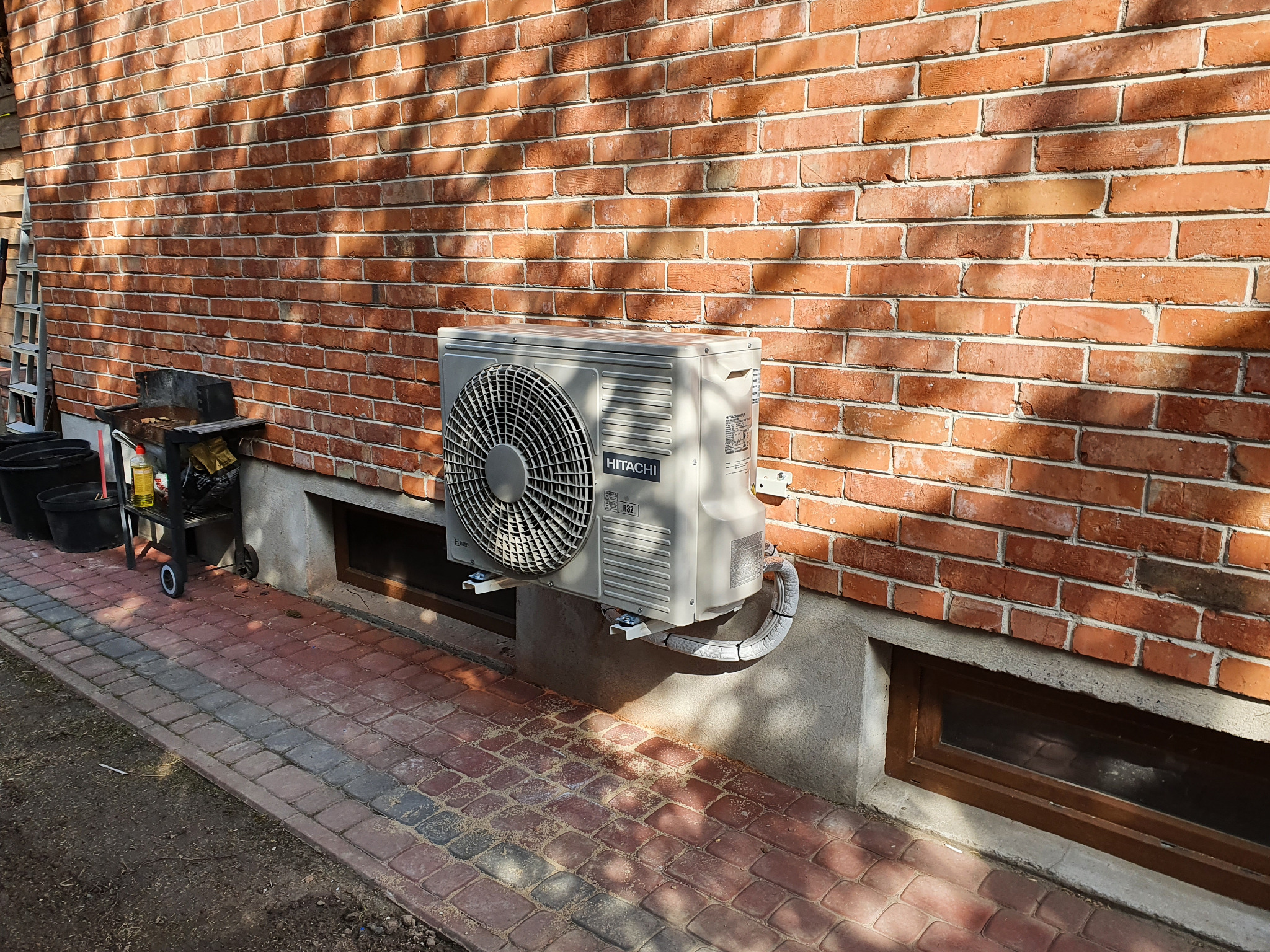 Zewnętrzna jednostka klimatyzacji Hitachi zamontowana na ceglanej ścianie budynku w Przedborzu, widoczne kable w osłonie i fragment chodnika z kostki brukowej.