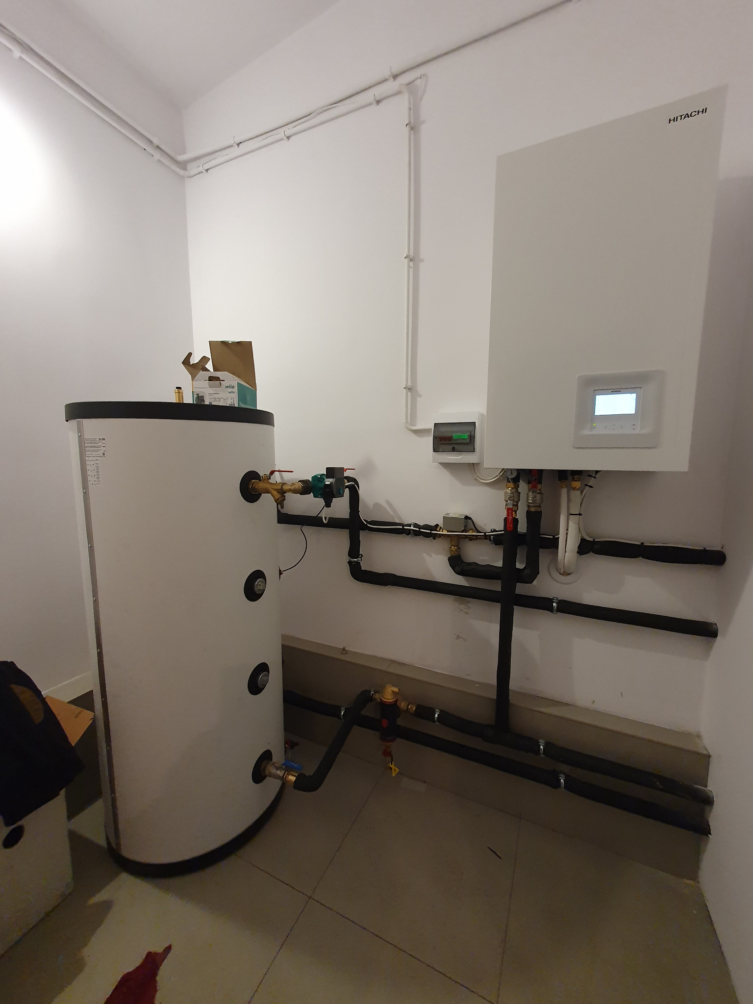Biała pompa ciepła Hitachi zamontowana na ścianie w pomieszczeniu technicznym, z podłączonymi czarnymi rurami i zbiornikiem buforowym, widoczna skrzynka elektryczna i elementy instalacji.