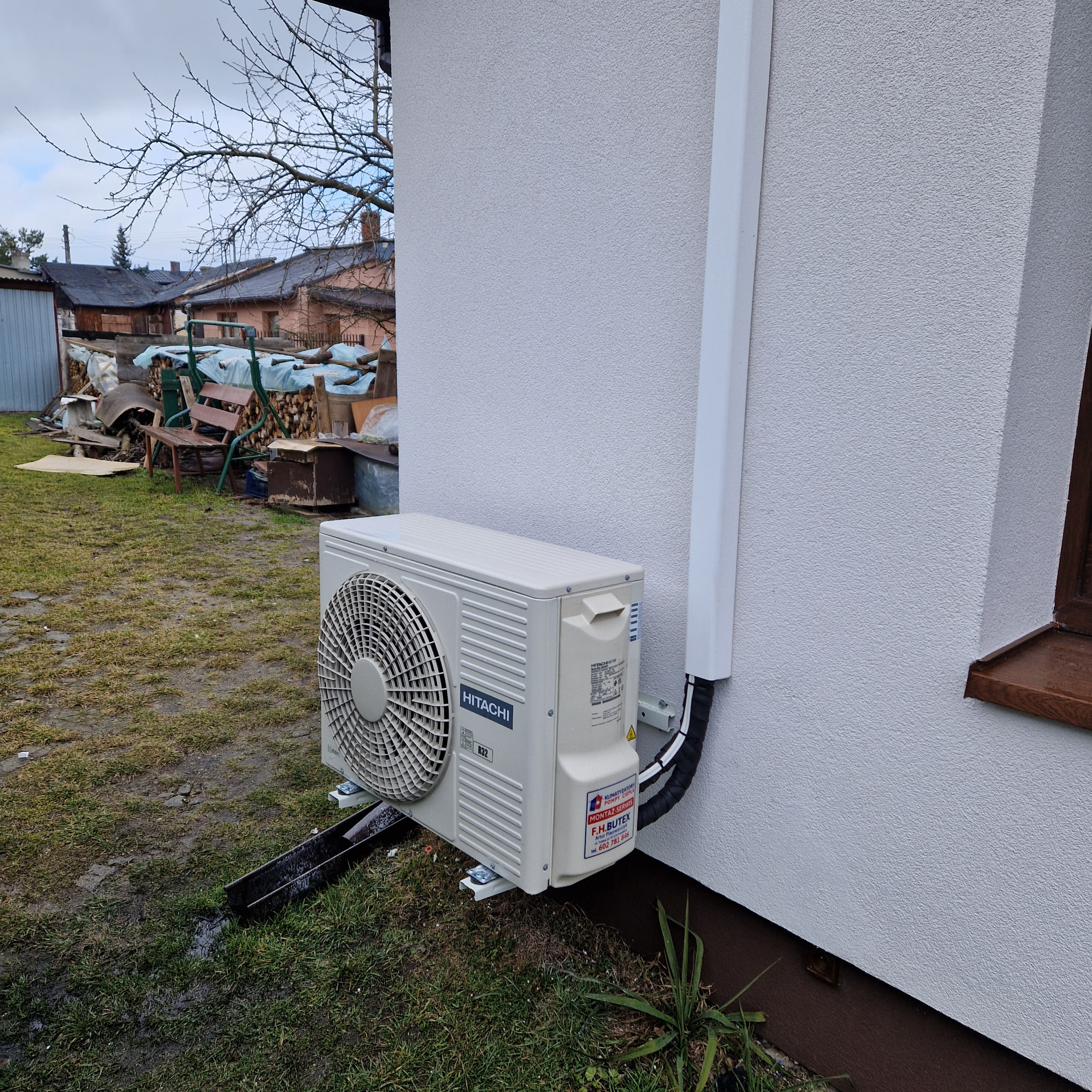 Zewnętrzna jednostka klimatyzacyjna Hitachi zamontowana na elewacji budynku z widoczną rurą odprowadzającą skropliny i osłoną przewodów. Na obudowie jednostki naklejka z danymi kontaktowymi firmy...