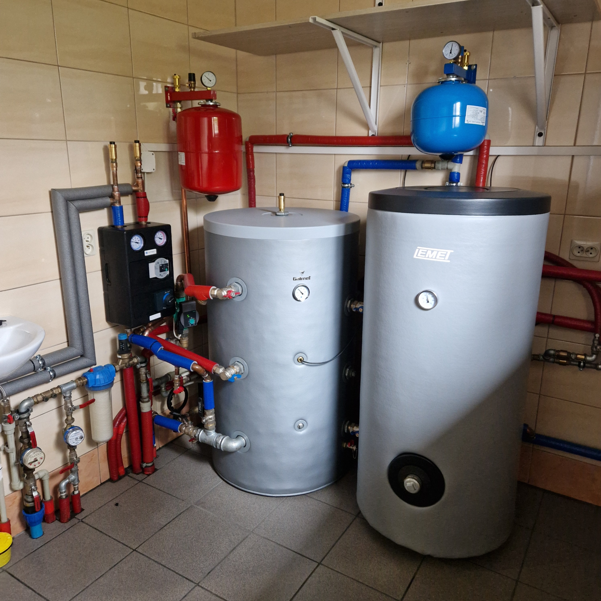 Dwa szare zbiorniki akumulacyjne ciepłej wody z podłączeniami rur w kolorach czerwonym i niebieskim, zainstalowane w kotłowni z beżowymi płytkami ceramicznymi na ścianach i podłodze.