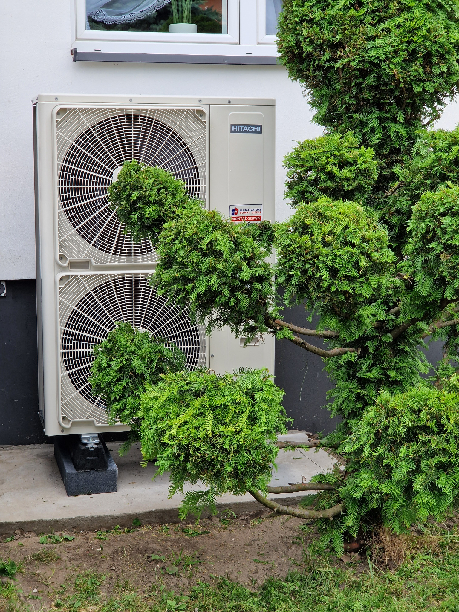 Zewnętrzna jednostka klimatyzacji Hitachi, z dwoma wentylatorami, zamontowana przy białej ścianie domu, częściowo zasłonięta ozdobnym krzewem.