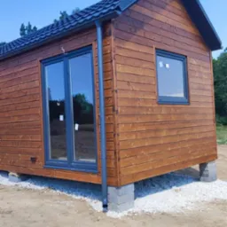 Mały, drewniany budynek z ciemną dachówką, posadowiony na betonowych bloczkach, z szarymi oknami i drzwiami przesuwnymi, otoczony ziemią i białym żwirem.