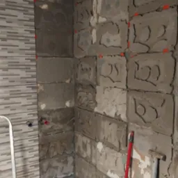 Ściana w trakcie układania płytek, widoczne nierówności i resztki zaprawy, fragment ściany z ułożonymi płytkami w odcieniach szarości, drabina i narzędzia glazurnicze w tle.