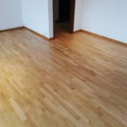 Jasne, nowo ułożone panele parkietowe w pustym pokoju z białymi ścianami i brązowymi listwami przypodłogowymi, widoczne drzwi do innego pomieszczenia.