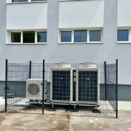 Energia Capital Sp z o.o. - Trzy zewnętrzne jednostki systemu grzewczo-chłodzącego, w tym dwie większe, zabezpieczone czarnym ogrodzeniem panelowym, na tle jasnoszarego budynku z białymi oknami.