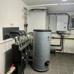 Energia Capital Sp z o.o. - Czysta kotłownia z szarym zbiornikiem ATRINNITY i jednostką Ecodan. Widoczny czarny rozdzielacz z rurami, zaworami i manometrami, na tle białych ścian i szarych płytek.