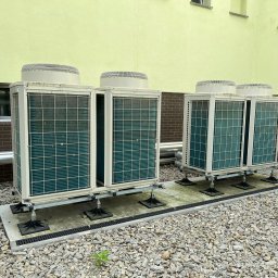 Energia Capital Sp z o.o. - Cztery zewnętrzne jednostki pomp ciepła, ustawione parami na betonowych podstawach z regulowanymi nóżkami, obok jasnożółtego budynku na żwirowym podłożu.