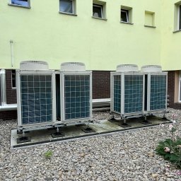Energia Capital Sp z o.o. - Cztery zewnętrzne jednostki grzewcze, zamontowane parami na podwyższonych platformach, na tle jasnożółtej elewacji z uchylonymi oknami. Podłoże z jasnego żwiru.