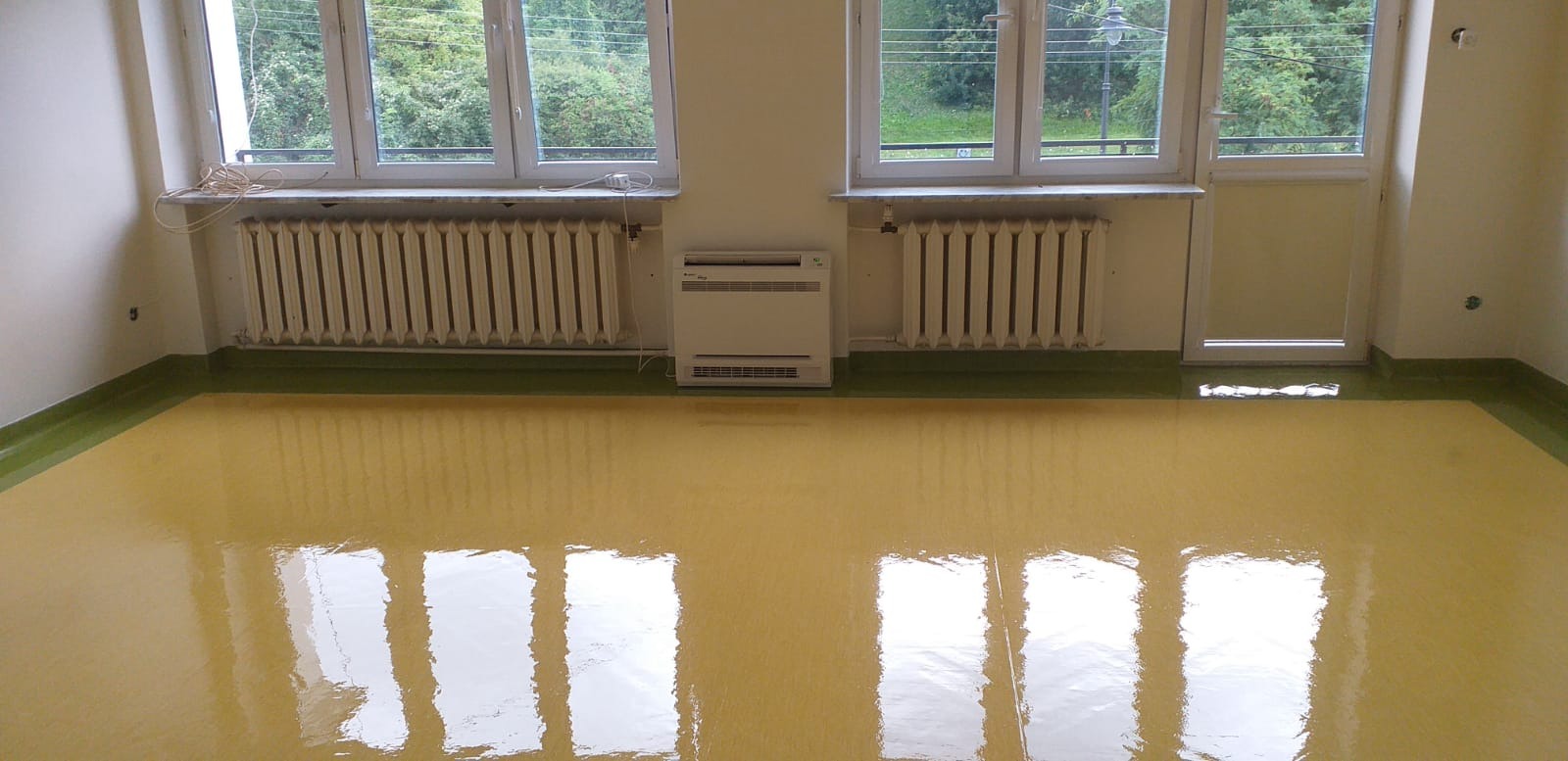 Świeżo wylana, połyskująca, żółta żywica epoksydowa na podłodze w jasnym pomieszczeniu z dwoma oknami i grzejnikami.