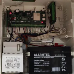 Otwarta obudowa centrali alarmowej z widoczną elektroniką, transformatorem i akumulatorem, okablowanie wewnętrzne.