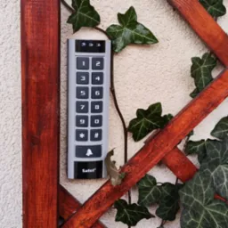 Klawiatura domofonu Satel zamontowana na elewacji z drewnianą kratką, opleciona bluszczem.