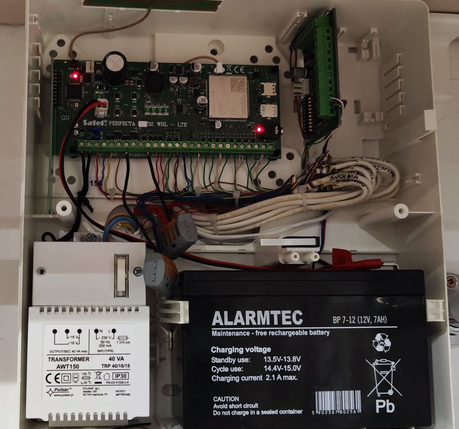 Otwarta obudowa centrali alarmowej z widoczną elektroniką, transformatorem i akumulatorem, okablowanie wewnętrzne.