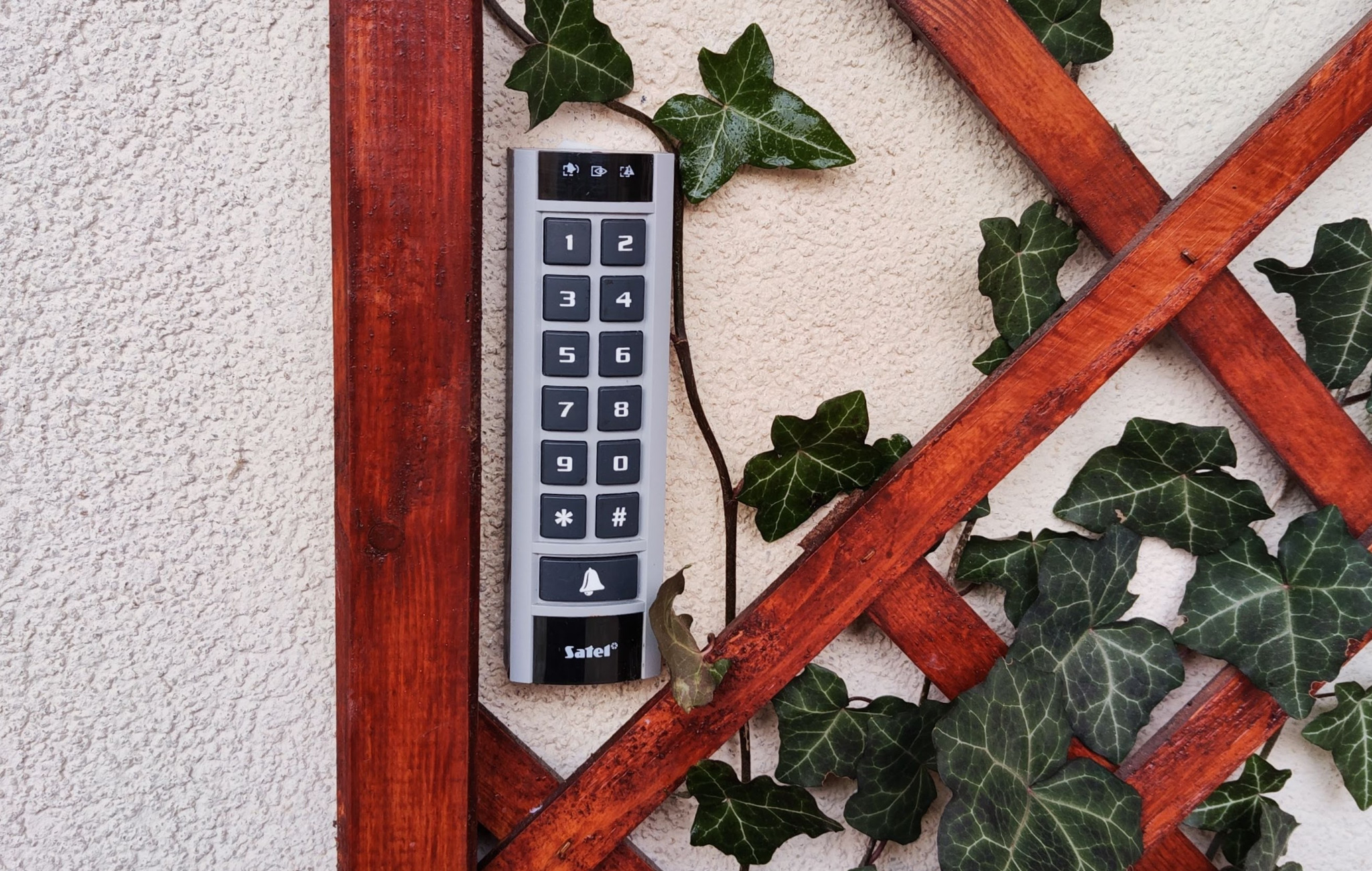 Klawiatura domofonu Satel zamontowana na elewacji z drewnianą kratką, opleciona bluszczem.