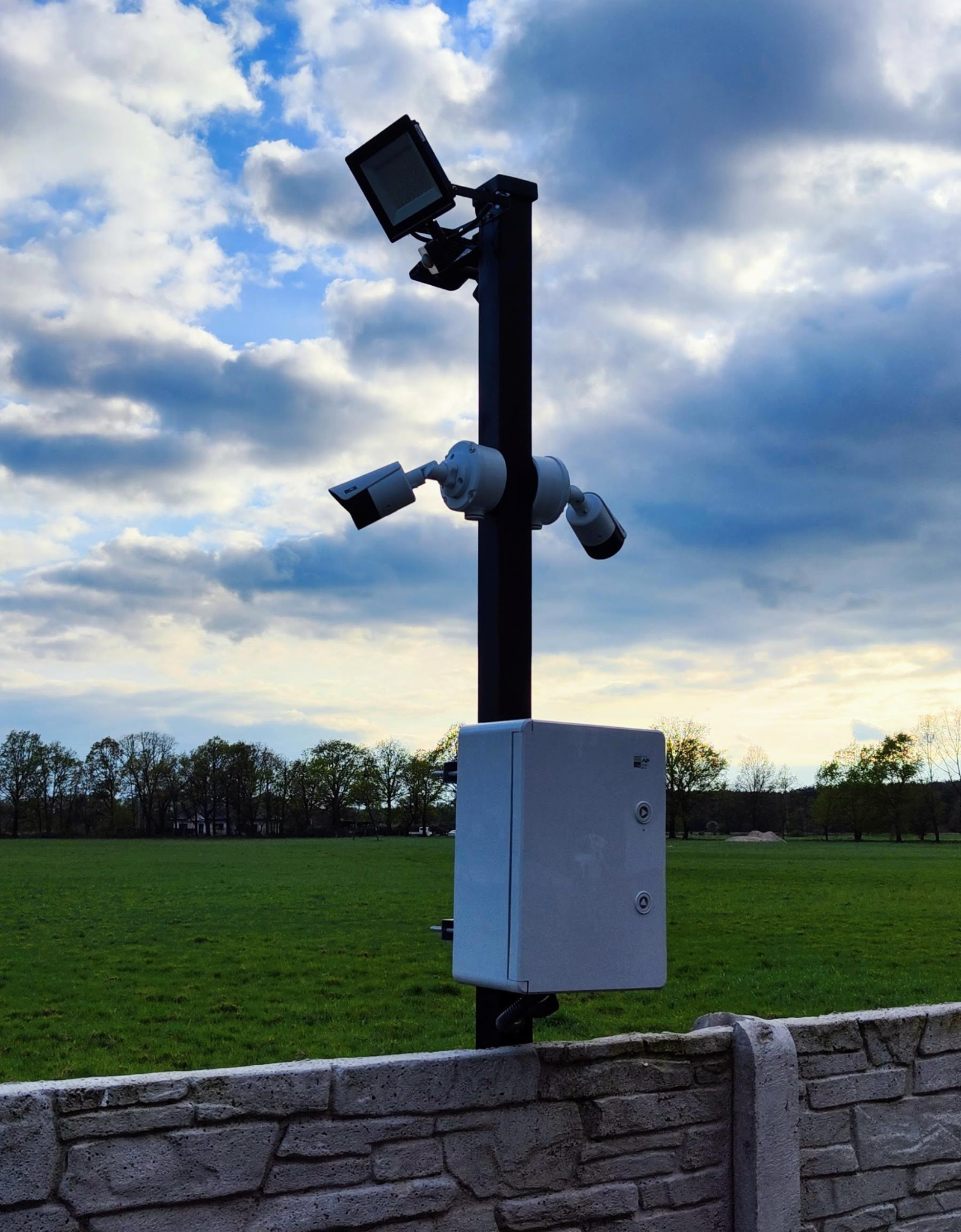 Słup z trzema kamerami monitoringu i oświetleniem LED, zamontowany za betonowym ogrodzeniem, na tle zielonego pola i pochmurnego nieba.