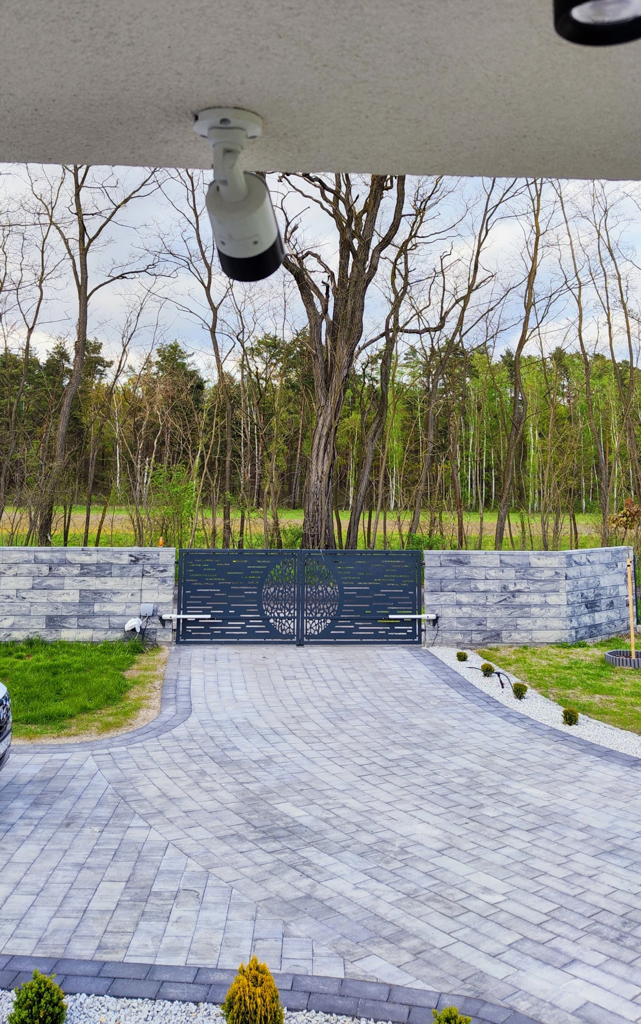 Nowoczesna automatyczna brama wjazdowa z dekoracyjnym wzorem na tle lasu, widoczna kamera monitoringu na podjeździe wyłożonym kostką brukową.