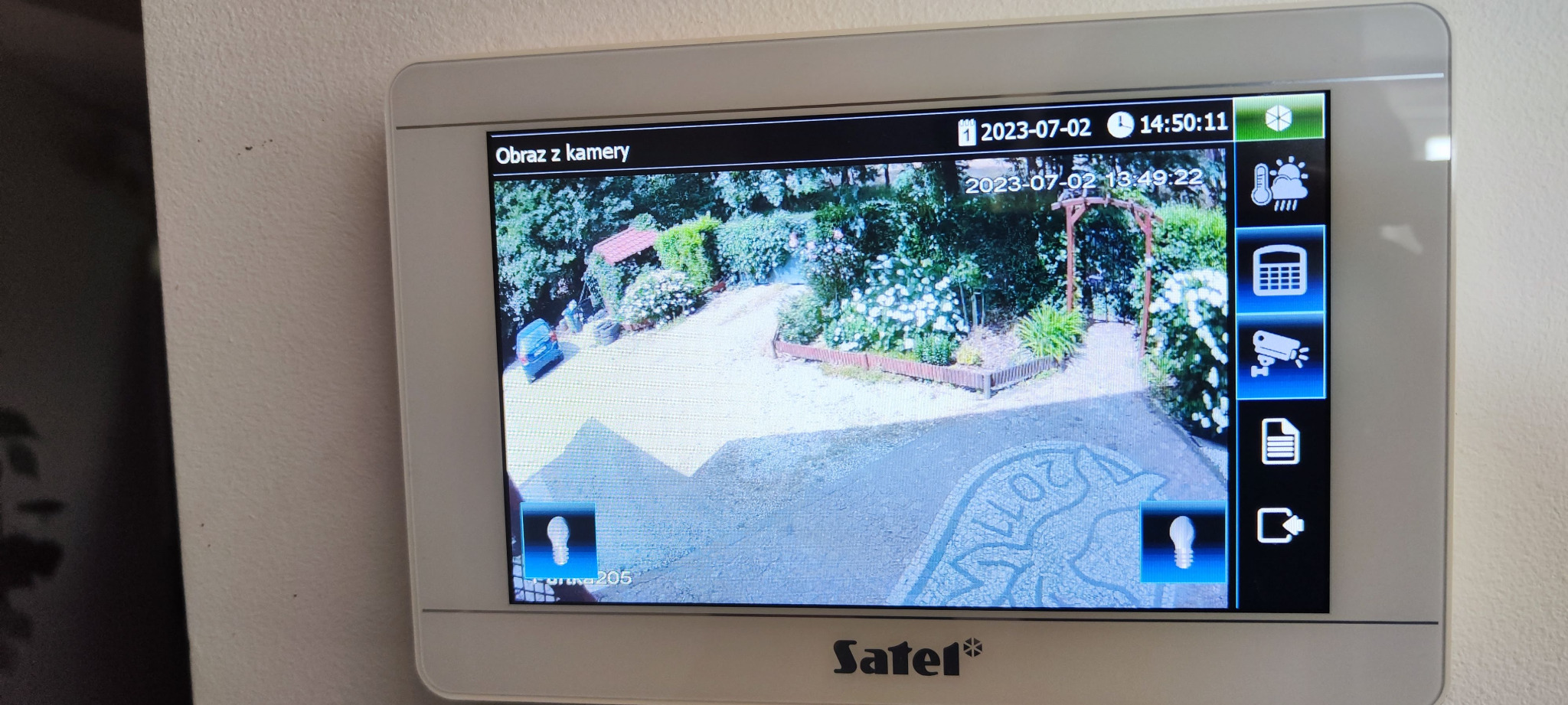 Ekran wideodomofonu marki Satel wyświetlający obraz z kamery: widok na podjazd z samochodem, rabatami kwiatowymi i drewnianą pergolą.