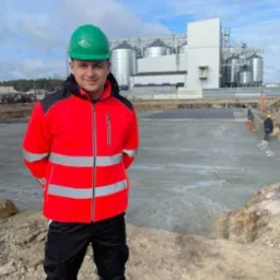 Mężczyzna w zielonym kasku i czerwonej kurtce roboczej na placu budowy z widocznymi silosami w tle.