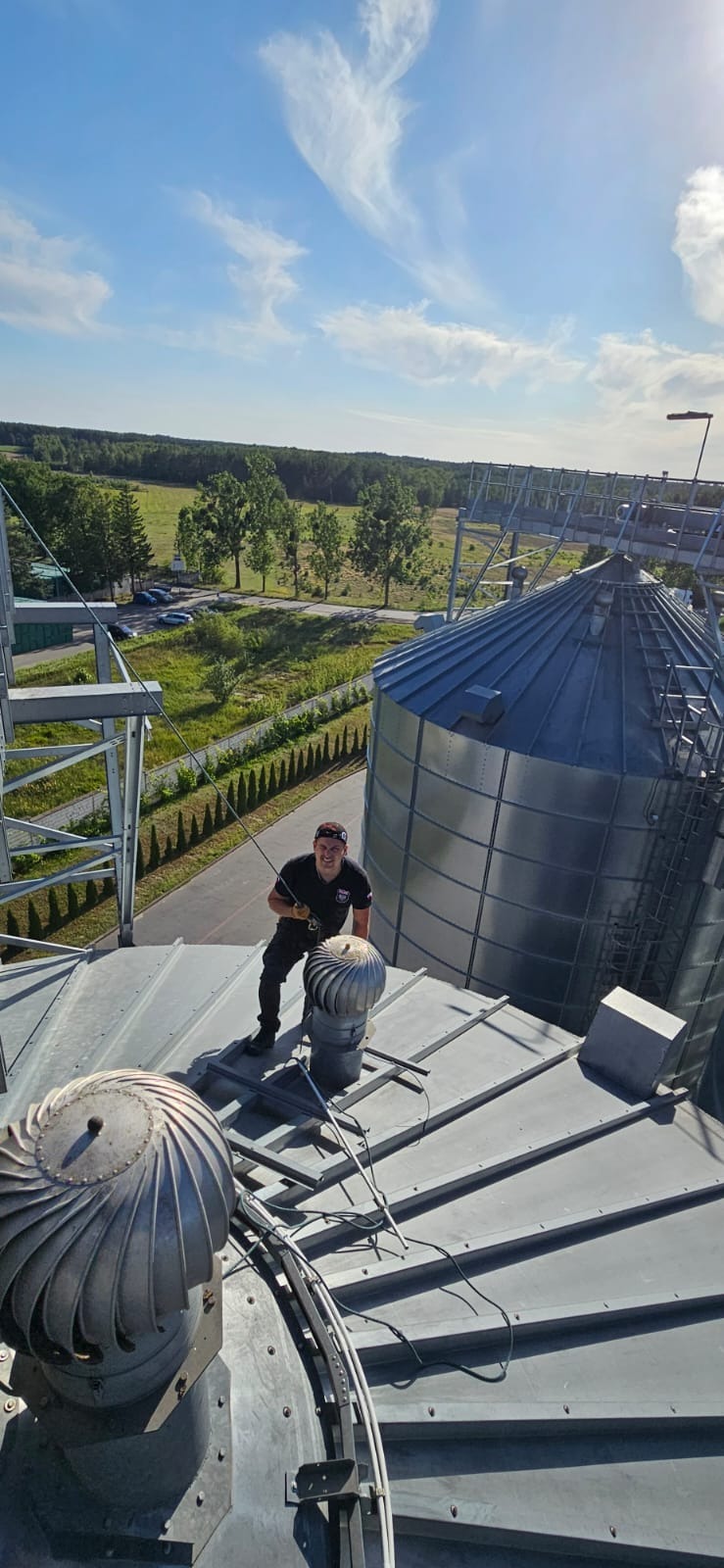 Pracownik na dachu silosu zbożowego podczas inspekcji lub prac konserwacyjnych. Widoczne elementy wentylacji. W tle krajobraz wiejski i błękitne niebo.