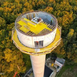 Widok z lotu ptaka na budowę cylindrycznej wieży z betonu, otoczonej lasem, z widocznym zbrojeniem i deskowaniem na g&oacute;rze oraz pracownikami w kaskach.