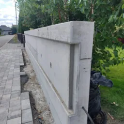 Nowo wybudowany, surowy, betonowy mur oporowy z widocznymi elementami konstrukcyjnymi, położony wzdłuż chodnika z kostki brukowej, z workiem na śmieci i przewodami u podstawy.