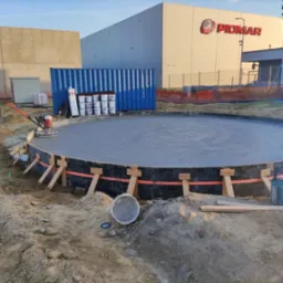 Okrągła, świeżo wylana płyta betonowa w szalunku, obrabiana zacieraczką mechaniczną. W tle kontener, palety z materiałami budowlanymi i budynek z logo Piomar.