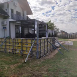 Żółte szalunki Doka z metalowymi wzmocnieniami przygotowane do wylania fundamentów przy nowoczesnym domu z balkonem i pergolą.