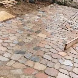 Nieregularna kamienna nawierzchnia w trakcie układania, widoczne mokre kamienie w odcieniach szarości, brązu i różu, z fragmentami ułożonymi w rzędach, obok stos płyt i drewniany element...