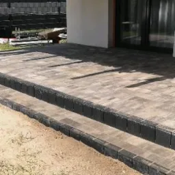 Wykończony taras z kostki brukowej w odcieniach szarości, z dwoma stopniami obłożonymi ciemnymi obrzeżami, widok z boku na tle jasnej elewacji budynku i zieleni w tle.