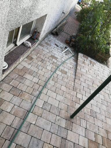 Ułożona kostka brukowa w odcieniach beżu i szarości, widoczny fragment ściany budynku z oknem, wąż ogrodowy i roślinność w tle.