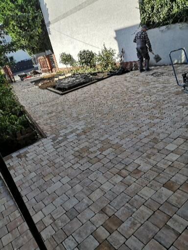 Ułożona kostka brukowa w odcieniach szarości i brązu, widoczny mężczyzna z łopatą i ubijarka w tle, prace wykończeniowe w ogrodzie.