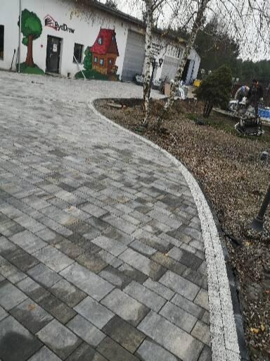 Świeżo ułożona, zakrzywiona ścieżka z szarej kostki brukowej w Koronowie, z widocznym obrzeżem i budynkiem w tle z ręcznie malowanymi grafikami.