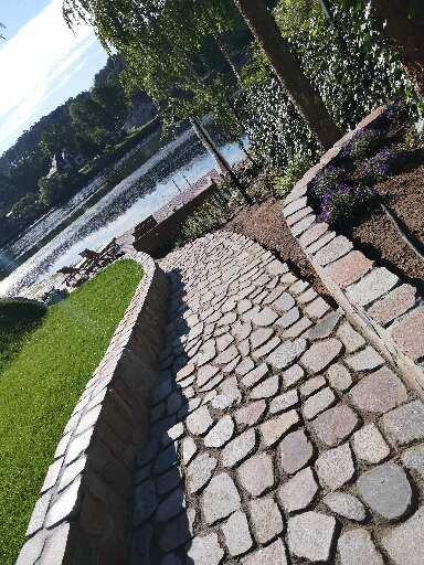 Ścieżka z nieregularnej kostki prowadząca w stronę jeziora, obok murku z kamienia i trawnika, w tle widoczne drzewa i woda.