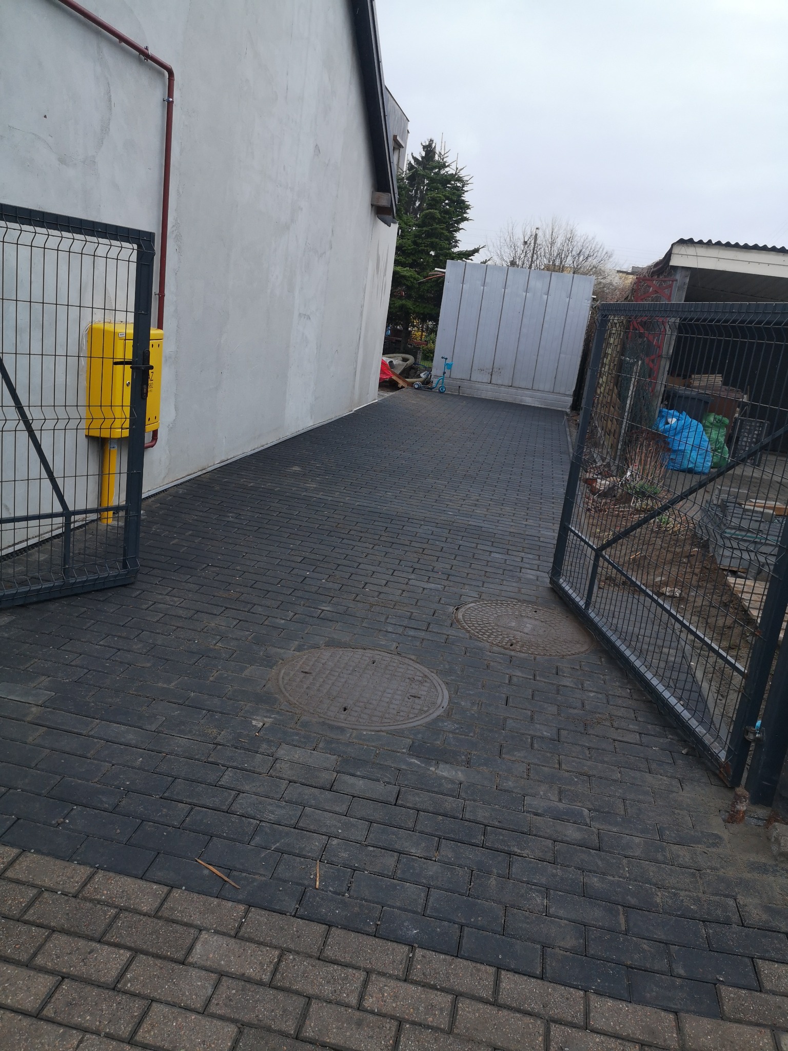 Ciemnoszara kostka brukowa ułożona wzdłuż ściany budynku, z widoczną skrzynką gazową i otwartą bramą panelową w tle.
