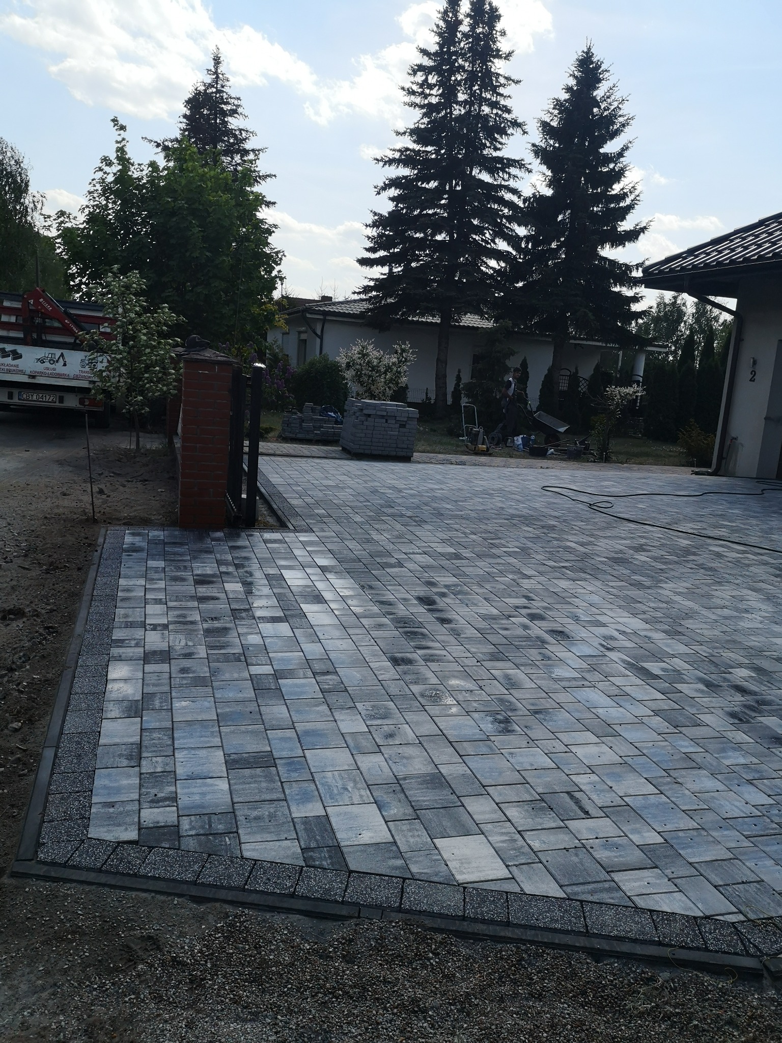 Nowo ułożony podjazd z szarej kostki brukowej, obrzeża z drobnego kamienia, brama wjazdowa z ceglanym słupkiem, w tle dom i wysokie drzewa.