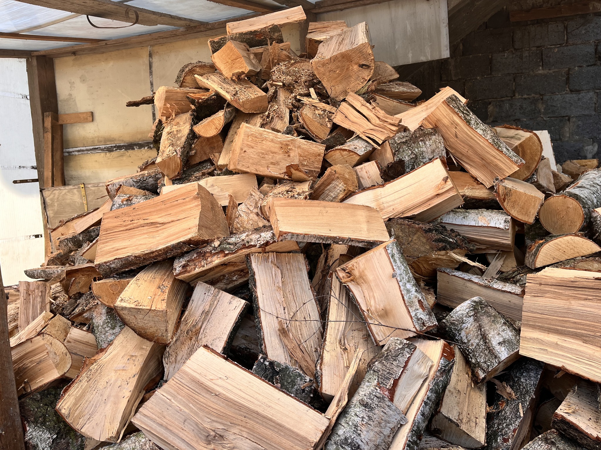 Stos pociętego drewna opałowego pod zadaszeniem, widoczne kawałki drewna z korą i bez kory, ułożone w nieuporządkowany sposób.