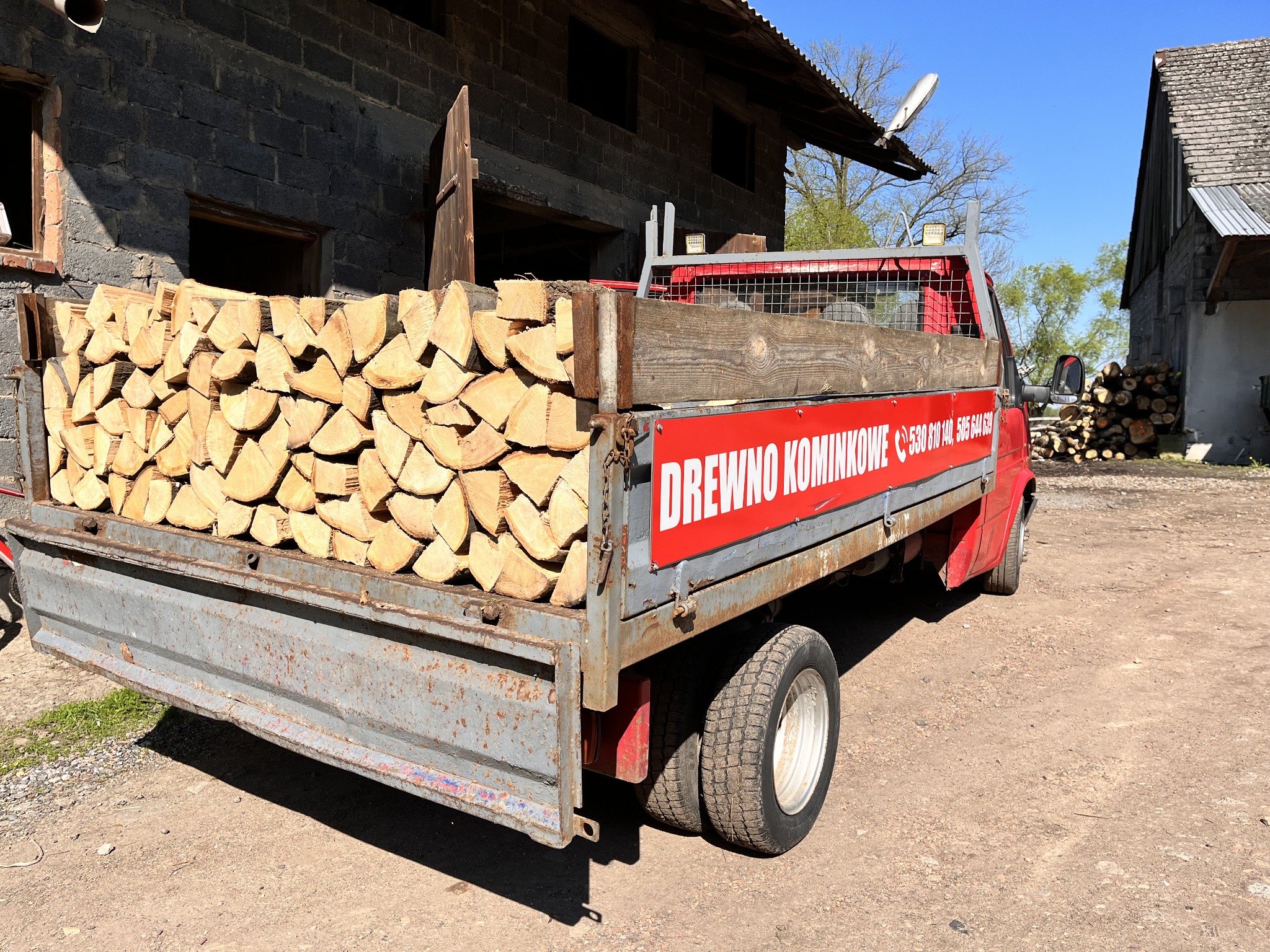 Ciężarówka z przyczepą wypełniona pociętymi kawałkami drewna na opał, z widocznym napisem reklamowym na burcie, zaparkowana na podwórzu przed budynkiem gospodarczym.