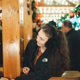 Fotografia Dawid Smułka - Uśmiechnięta młoda kobieta w nausznikach z futerkiem, oparta o drewnianą konstrukcję z metalowymi okuciami, w tle rozmyte dekoracje świąteczne.