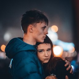 Fotografia Dawid Smułka - Młoda para w objęciach na tle miejskich świateł. Chłopak w niebieskiej kurtce, dziewczyna z wyrazistym makijażem, patrzy w obiektyw. Portret w miejskiej scenerii, nocne ujęcie.