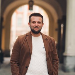 Fotografia Dawid Smułka - Mężczyzna z brodą w brązowym płaszczu i białym swetrze pozuje na tle łukowatego przejścia. Uśmiech, ręce w kieszeniach, swobodna elegancja.